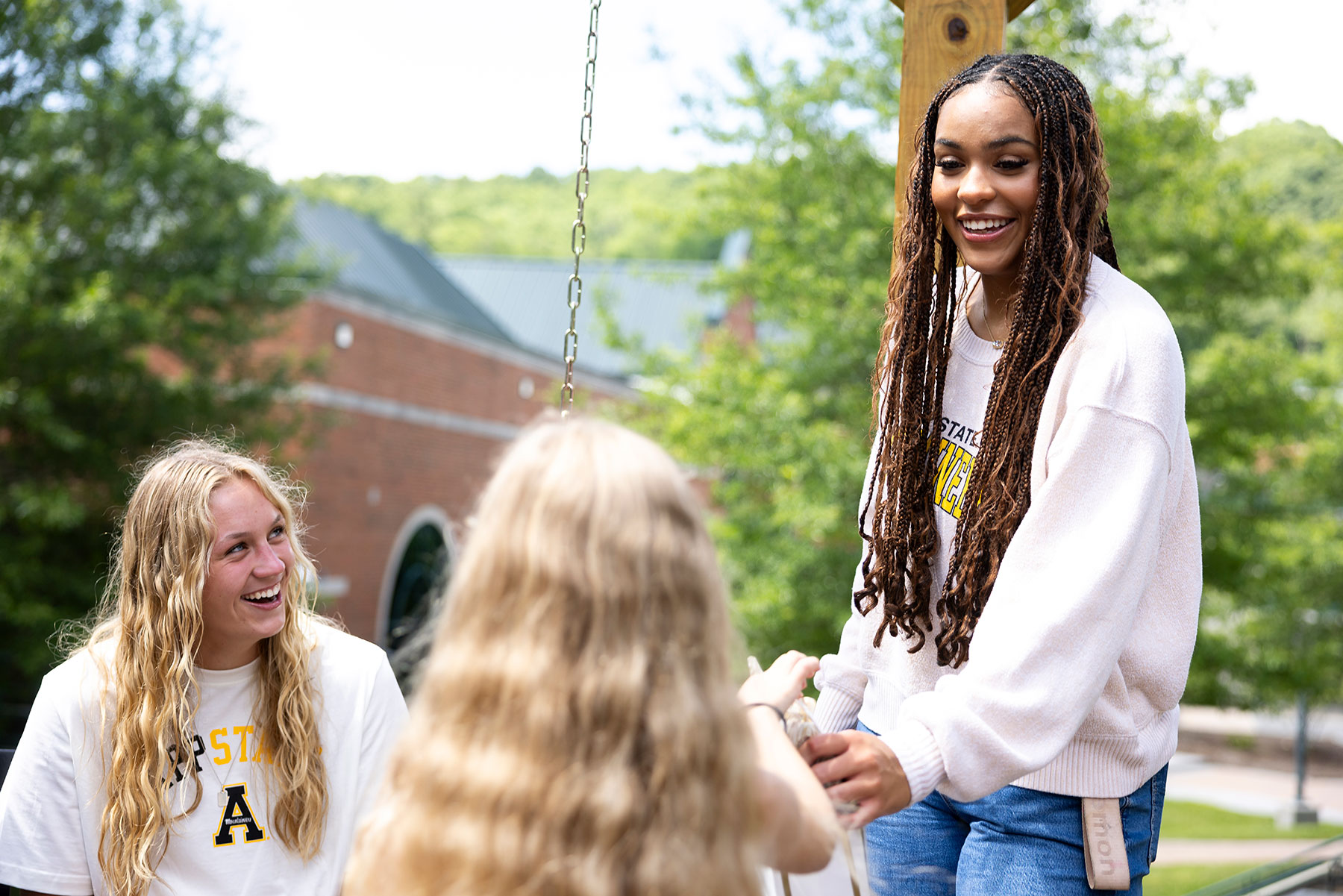 Three students smile and talk together outdoors on campus, wearing App State clothing.