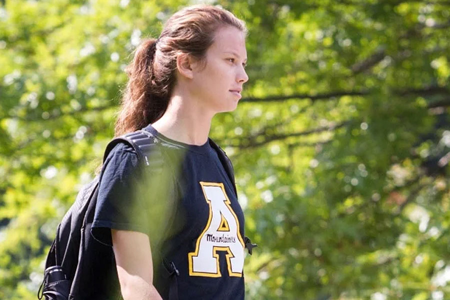 Student walking along a tree-lined campus pathway wearing an Appalachian State shirt and backpack.
