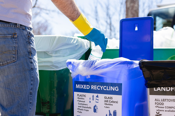 Gloved hand places a plastic bottle into a mixed recycling bin beside waste collection containers.
