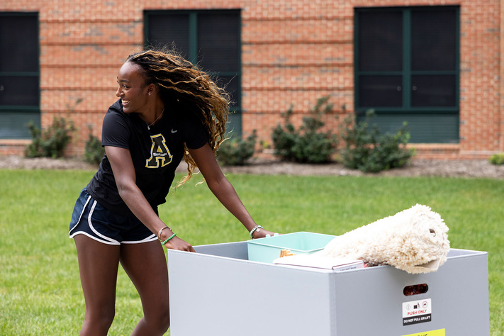 Student pushes a move-in cart filled with belongings across campus lawn near a residence hall.