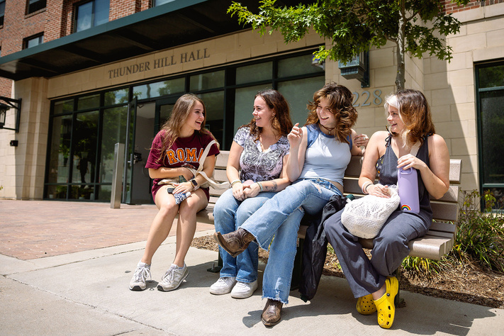 Four students laugh together on a bench outside the main entrance to Thunder Hill Hall.