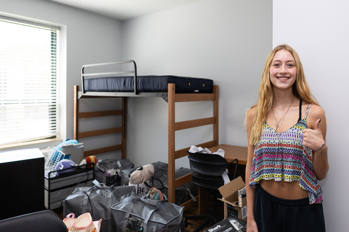 Student unpacking belongings in a residence hall room with lofted bed, desk, chair and move-in supplies.