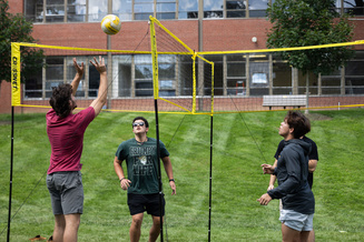 Four people play four-way volleyball on a grassy lawn outside a brick building.